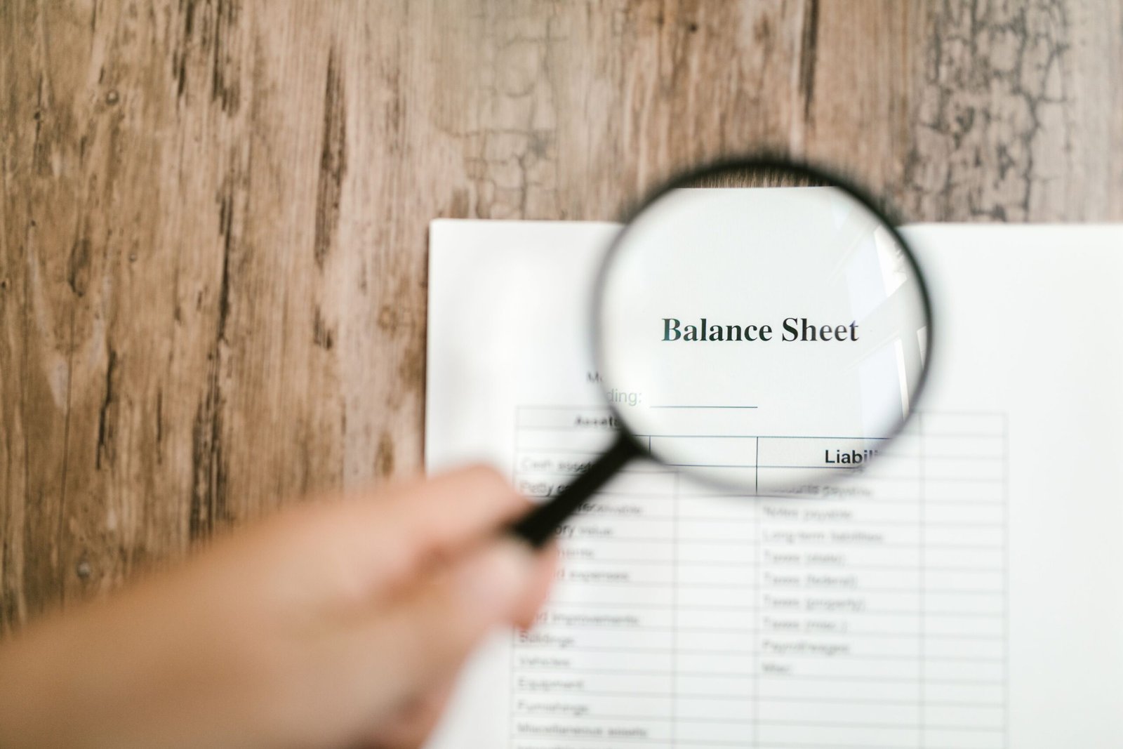 Close-up of a balance sheet document on wooden surface with a magnifying glass held by a hand.